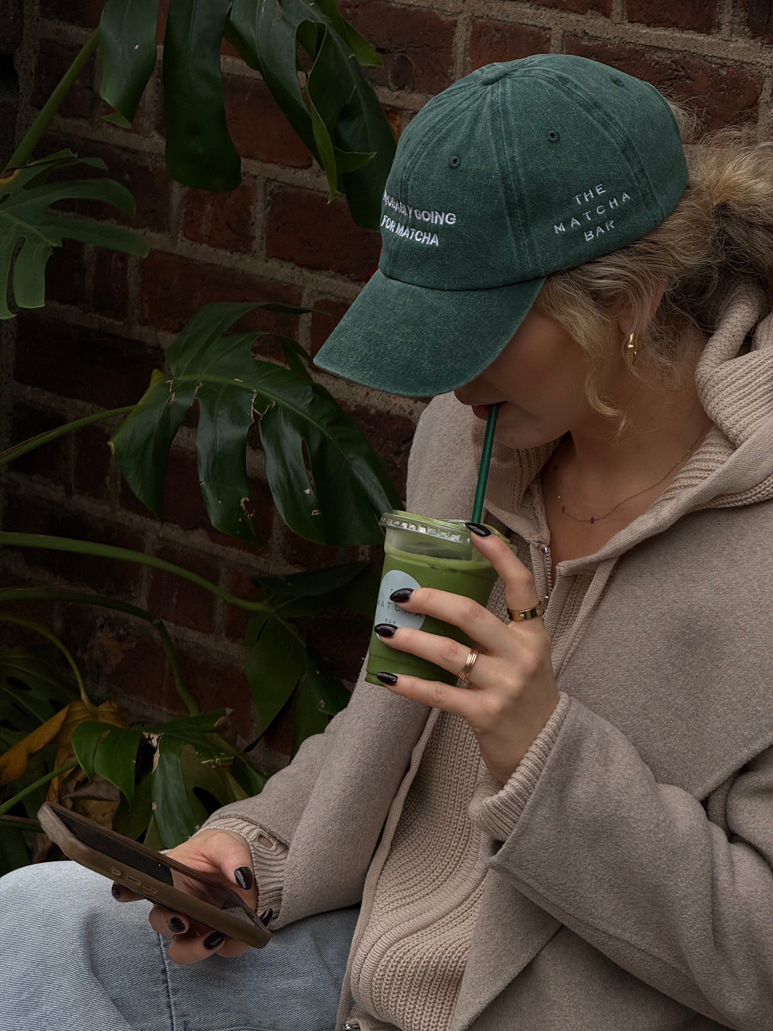 Person wearing a green cap and beige coat, holding a phone and a green drink against a brick wall with plants.